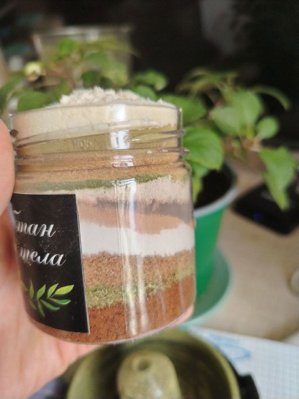 Пластиковая баночка с послойными порошками убтана: разноцветные глины, травы и специи, в руках на фоне растений.