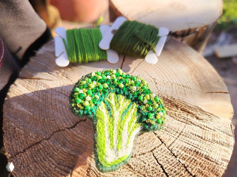 Брошь «брокколи» на пеньке рядом с катушками ниток и материалами для вышивки; подчёркнута идея ручной работы и процесса изготовления.