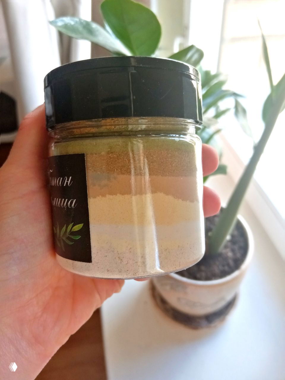 Стеклянная баночка в руке у окна: видны многослойные порошковые смеси (убтаны/травяные порошки) на фоне горшка с растением и подоконника.