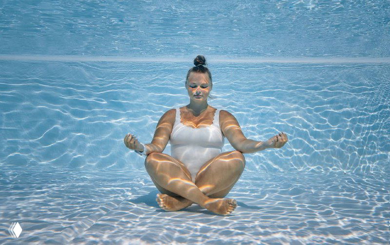 Женщина в белом купальнике погружена под воду в бассейне в позе лотоса, расслабленно медитирует и контактирует с водой.