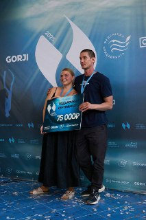 Двое людей позируют с большим сертификатом на фоне баннера фестиваля, видны медали на шее и праздничная атмосфера.