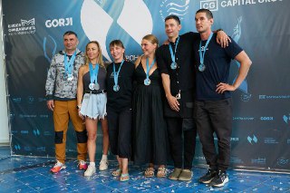 Группа фридайверов и организаторов на фоне брендового фона, все в медалях и с улыбками после церемонии награждения.
