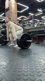 Человек в спортивной одежде выполняет становую тягу с тяжелой штангой в современном тренажерном зале, виден резиновый пол