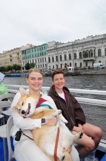 Две женщины на палубе кораблика, одна держит корги в руках; на заднем плане набережная с домами, летняя прогулка.