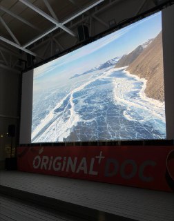 Фото зала кинофестиваля: крупный проекционный экран показывает кадр с замёрзшим морем и ледяной поверхностью у берега; внизу виден баннер ORIGINAL+ DOC.