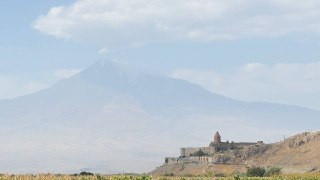 Панорамный вид на гору Арарат с равнины; на переднем плане видна каменная церковь или монастырь и лёгкие облака.