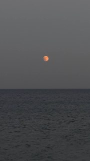 Фотография оранжевой Луны над тёмным морем во время лунного затмения, минимальный горизонт и спокойная водная поверхность.