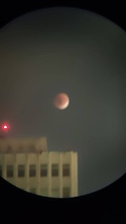 Крупный план Луны через оптический прибор, размытый план с городской архитектурой и тёплым свечением в кадре.