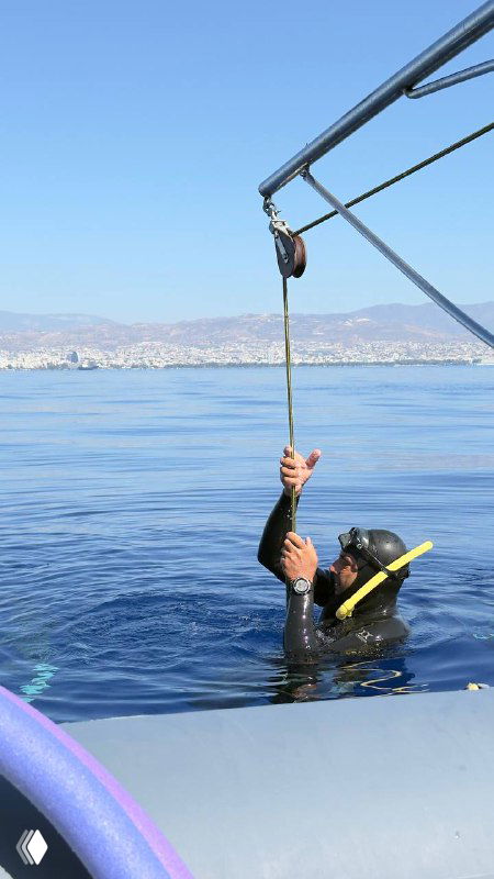 Ныряльщик на поверхности держится за трос рядом с лодкой, подготовка к глубокому или тренировочному погружению на фоне спокойного моря и побережья.