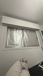 Окно в комнате, заклеенное алюминиевой фольгой для блокировки света; видна часть стены, подоконник и угол кровати, создавая затемнённую обстановку в спальне.