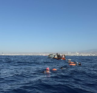 Группа фридайверов в открытом море рядом с поддерживающими лодками; видна линия берега, несколько буйков и спокойная ясная погода.