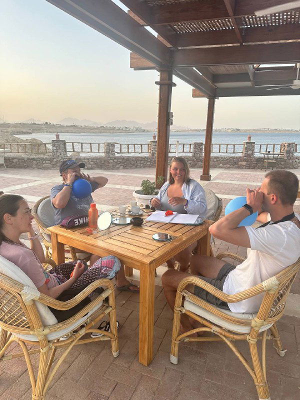 Группа фридайверов за деревянным столом на открытой веранде тренируется с носодуем: несколько человек надувают резиновые баллоны перед занятием, виден берег