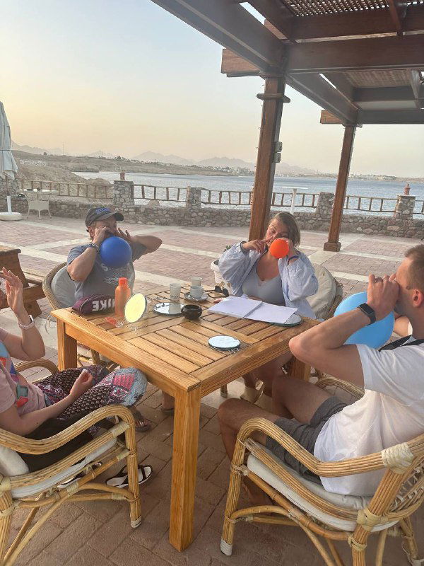 Участники упражнения пробуют носодуй: синий и оранжевый баллоны в руках, тренинг проходит у моря на открытой веранде с видом на залив и прогулочную набережную