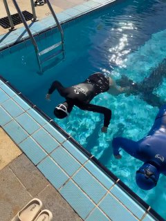 Фридайвер в гидрокостюме лежит лицом в воду у бортика открытого бассейна во время лагерной тренировки, видны лестница и плитка.