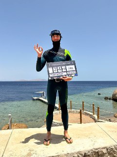Фридайвер в гидрокостюме и маске держит табличку с результатом, показывает жест "ок" на фоне моря и причала