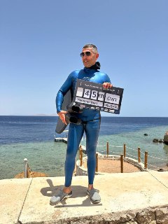 Мужчина в гидрокостюме стоит на каменном берегу, держит табличку с отметкой 45 м и снаряжение, позирует на фоне моря