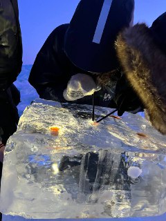 Человек в теплой одежде сверлит ледяной блок на берегу Байкала, видно инструменты и кусок прозрачного льда в руках группы.