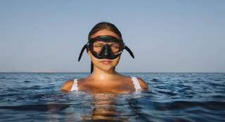 Человек в маске для ныряния, погружённый по шею в море; видна спокойная поверхность воды, ровный горизонт и безоблачное небо.