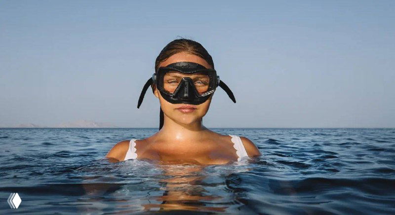 Фридайвер в маске, выглядывающий из воды на спокойном море; кадр подчёркивает тему ныряний, поездок и летних тренировок.