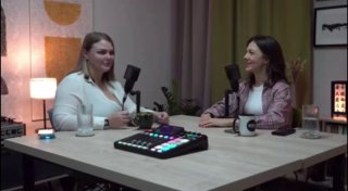Две женщины в студии подкаста сидят за столом с микрофонами, микшером и кружками; разговор в уютной акустической студии во время записи.