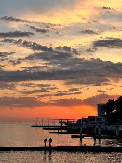 Закат над морским причалом в Сочи: оранжево-красное небо, силуэты людей и причалов, спокойная прибрежная атмосфера на побережье.