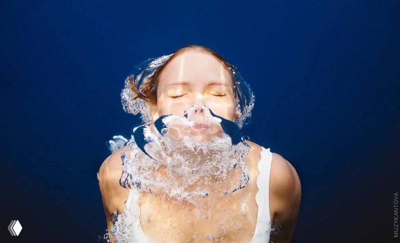 Женщина на крупном плане выныривает из воды, лицо и губы над поверхностью, вокруг брызги и пузыри — образ, подчёркивающий тему дыхания и фридайвинга.