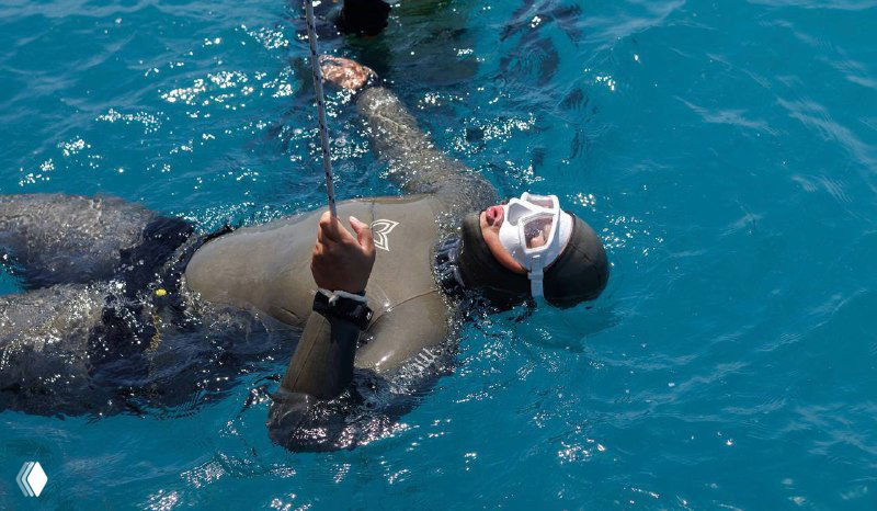 Фридайвер на поверхности воды держится за страховочный буй, видна маска и дыхательная трубка, спокойное море вокруг.
