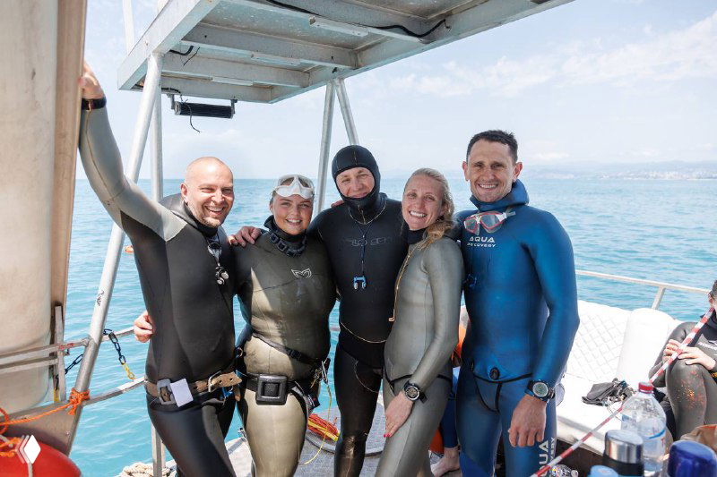 Группа участников в гидрокостюмах стоит вместе на палубе и улыбается: командный дух и поддержка во время чемпионата в Сочи.