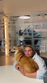 Девушка в белой одежде держит коричневого пса на руках в светлом офисном пространстве, видна мебель и стеклянная перегородка.