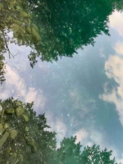 Крупный план берега и дна озера: чистая вода, видны камни и отражения облаков и прибрежной растительности в воде.