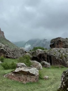 Горный пейзаж под плотными низкими тучами: скалы, влажная зелёная долина и туман, стелющийся между вершинами и уступами.