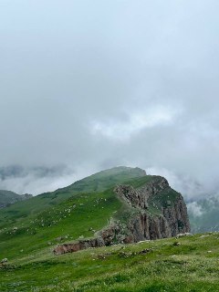 Зелёный горный хребет с обширными лугами и туманом, видны скалы и крутые склоны, кадр передаёт влажную прохладу места.