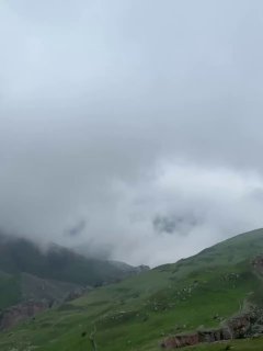 Туманное горное поле с зелёными лугами и размытыми скалами, низкие облака закрывают вершины, создавая прохладную атмосферу.