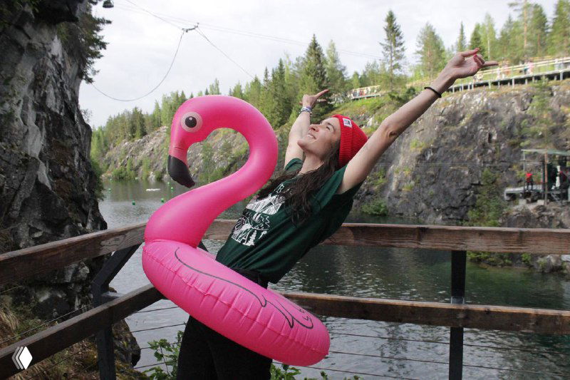 Девушка с надувным розовым фламинго на деревянной площадке у Мраморного карьера Рускеала, вокруг видны высокие скалы и вода, летняя атмосфера.