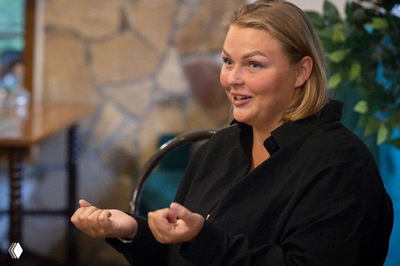 Женщина в тёмной одежде жестами показывает технику дыхания и продувки на занятии в помещении с нейтральным фоном