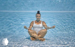 Женщина в белом купальнике под водой в бассейне в позе медитации; световые блики на коже, ощущение спокойного погружения.