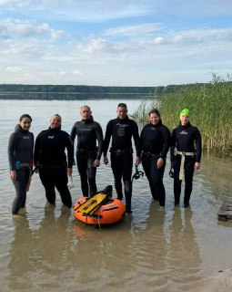 Группа фридайверов в гидрокостюмах стоит в мелкой воде у голубого озера с оранжевым буйком, камышами на берегу и лесом вдали.