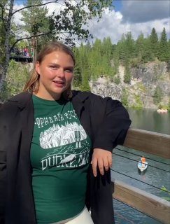 Женщина оперлась на деревянное ограждение у карьера с водой, на заднем плане отвесные скалы, хвойный лес и небо.