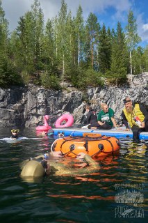 Группа людей у воды с надувными кругами, буйками и снаряжением для фридайвинга; сцена у поверхности с участниками и помощниками.