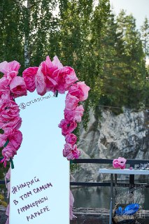 Декор фестиваля у воды: зеркало в розовой цветочной гирлянде на фоне скал, элемент оформления и атмосферная деталь мероприятия.
