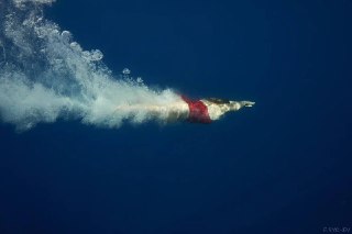 Фридайвер погружается в глубокую синюю воду, виден профиль ныряющего и длинный след пузырьков, создающий контекст техники.