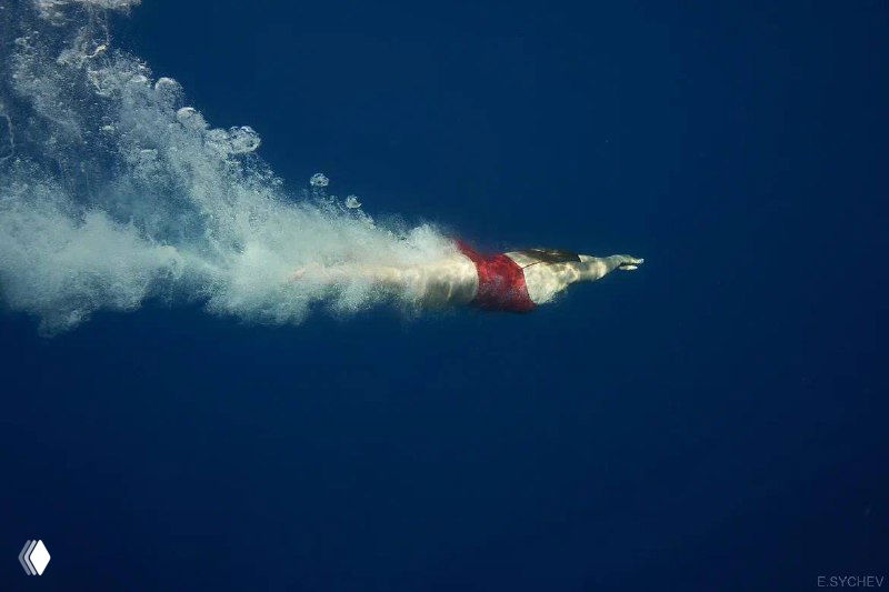 Подводный крупный план фридайвера в красном костюме, идущего вниз с пузырьковой струёй на фоне тёмно‑синей воды; логотип в углу.
