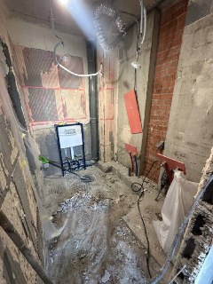 Комната в процессе ремонта: обнажённые стены, снятая отделка, строительный мусор на полу, видны трубы и коммуникации вокруг.