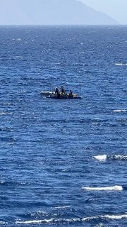 Небольшая вспомогательная лодка на открытой воде, люди на борту, видна поверхность моря с рябью, удалённый берег и горы вдалеке.