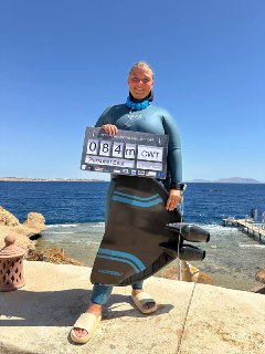 Автор в гидрокостюме стоит на каменном причале у моря, держит табличку с результатом и улыбается на фоне ясного синего неба.