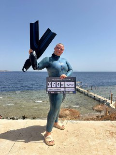 Человек в гидрокостюме стоит на берегу, держит ласты и табличку, на фоне виден пирс и спокойное море; портрет после погружения.
