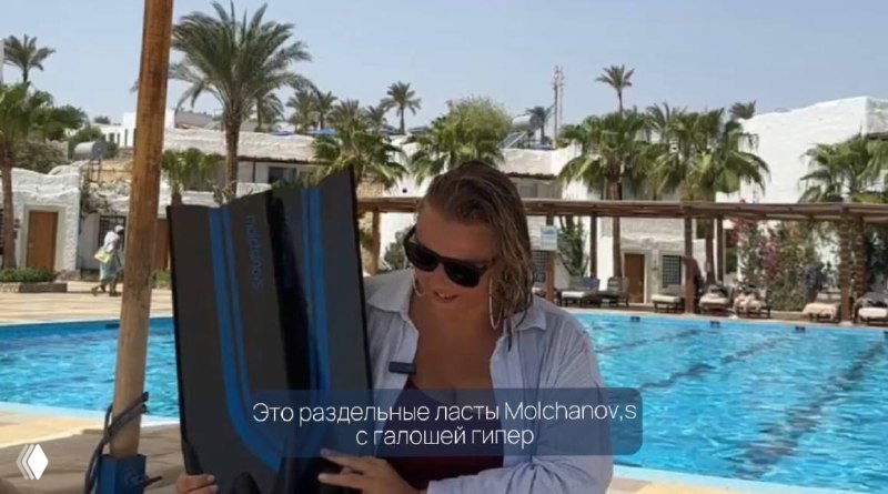 Автор (Ольга Маркина) сидит у бассейна в отеле и держит ласты Molchanovs; кадр из видеообзора с субтитрами на экране.