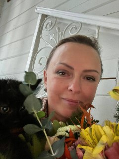 Улыбающаяся женщина с букетом жёлтых цветов, рядом частично видна чёрная кошка; на заднем плане видна белая решётка.