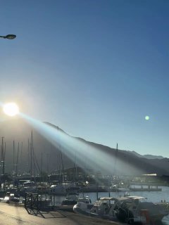 Причал с пришвартованными лодками на фоне гор, яркое солнце с лучом над водой, гавань с мачтами и спокойной поверхностью