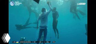 Подводный кадр соревнований по фридайвингу: спортсменка всплывает к поверхности, рядом судьи и участники на фоне синей воды, кадр из видео.